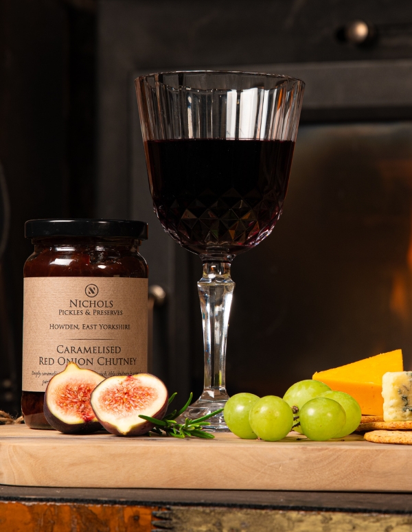 A cheeseboard with a jar of Nichols Pickles & Preserves Caramelised Red Onion Chutney. Also on the board is a goblet of red wine, grapes, figs, cheese and crackers.