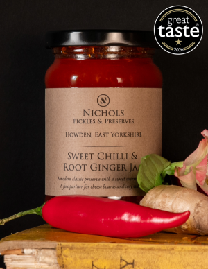 A jar of award winning sweet chilli and root ginger jam from Nichols Pickles & Preserves. Surrounding the jar is a red chilli, a piece of ginger and a flower.