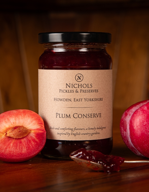 A jar of plum conserve from Nichols Pickles & Preserves on a wooden surface, surrounded by a whole plum and half a plum. In the foreground is a teaspoon with plum conserve on it.