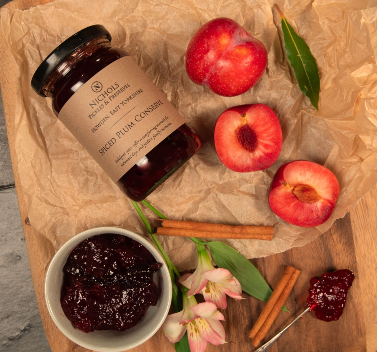Jar of Nichols Spiced Plum Conserve surrounded by fresh plums and cinnamon sticks.